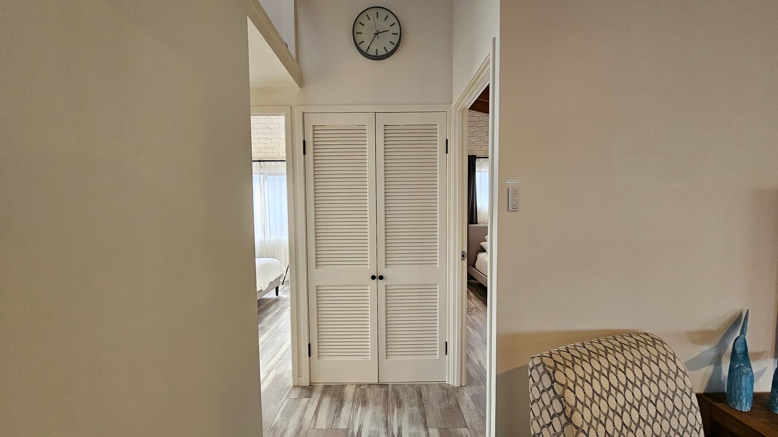 On the left, there is a doorway leading to a room with a bed and a large window with sheer curtains. In the middle, there are white louvred double doors with black handles and a wall clock above them. On the right, there is another doorway leading to a room with a bed and a large window with sheer curtains. In the foreground, there is a patterned armchair and a small table with a blue vase. The floor has laminate with a wooden design, and the ceiling is white with a recessed section.