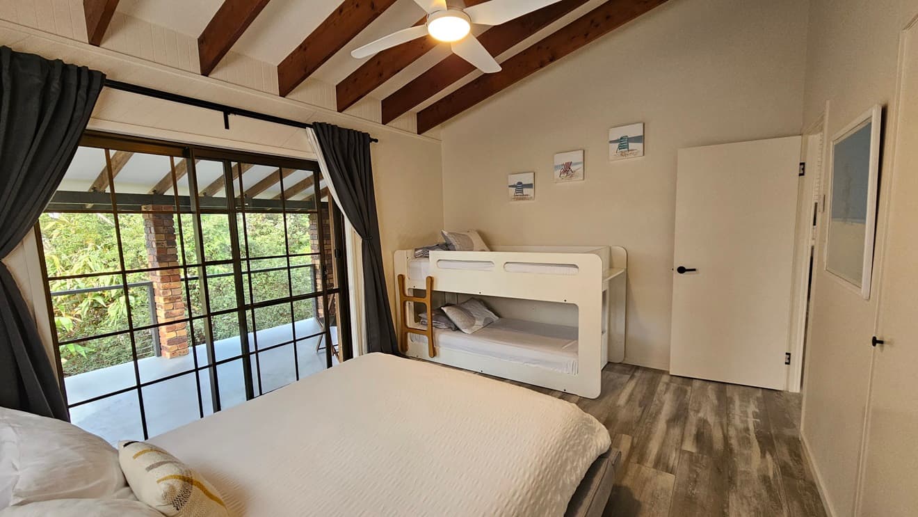 On the left, there is a large sliding glass door with black frames leading to a balcony with a view of greenery, covered by dark curtains. In the middle, there is a white bed with white bedding and a yellow cushion, positioned against a wall with two framed pictures above. On the right, there is a white bunk bed with grey bedding and a small wooden ladder, next to a white door. The floor has laminate with a wooden design and the ceiling has timber slats and a ceiling fan.