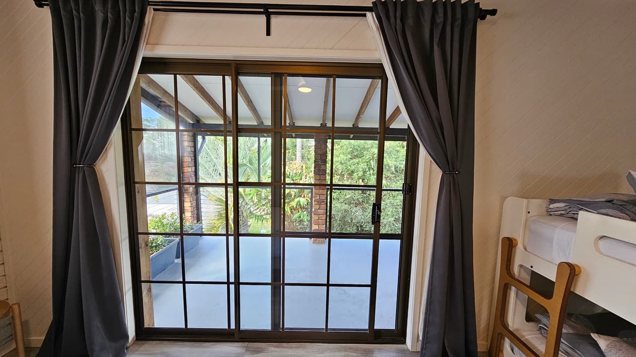 On the left, there is a white wall with a small section of a doorway visible. In the middle, there is a large sliding glass door with black frames, leading to a balcony with a view of greenery and palm trees, covered by dark curtains. On the right, there is a white bunk bed with grey bedding and a wooden ladder. The floor has laminate with a wooden design, and the ceiling has exposed wooden beams with recessed lighting.