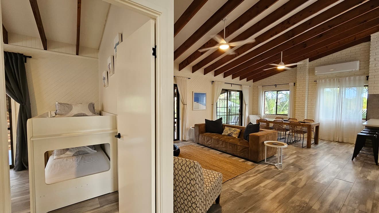 On the left, there is a doorway leading to a room with a white bunk bed and a large window with sheer curtains. In the middle, there are white louvred double doors and a wall clock above them. On the right, there is another doorway leading to a room with a bed and a large window with sheer curtains. In the foreground, there is a patterned armchair and a small table with a vase. The floor has laminate with a wooden design, and the ceiling is white with a recessed section and exposed wooden beams.