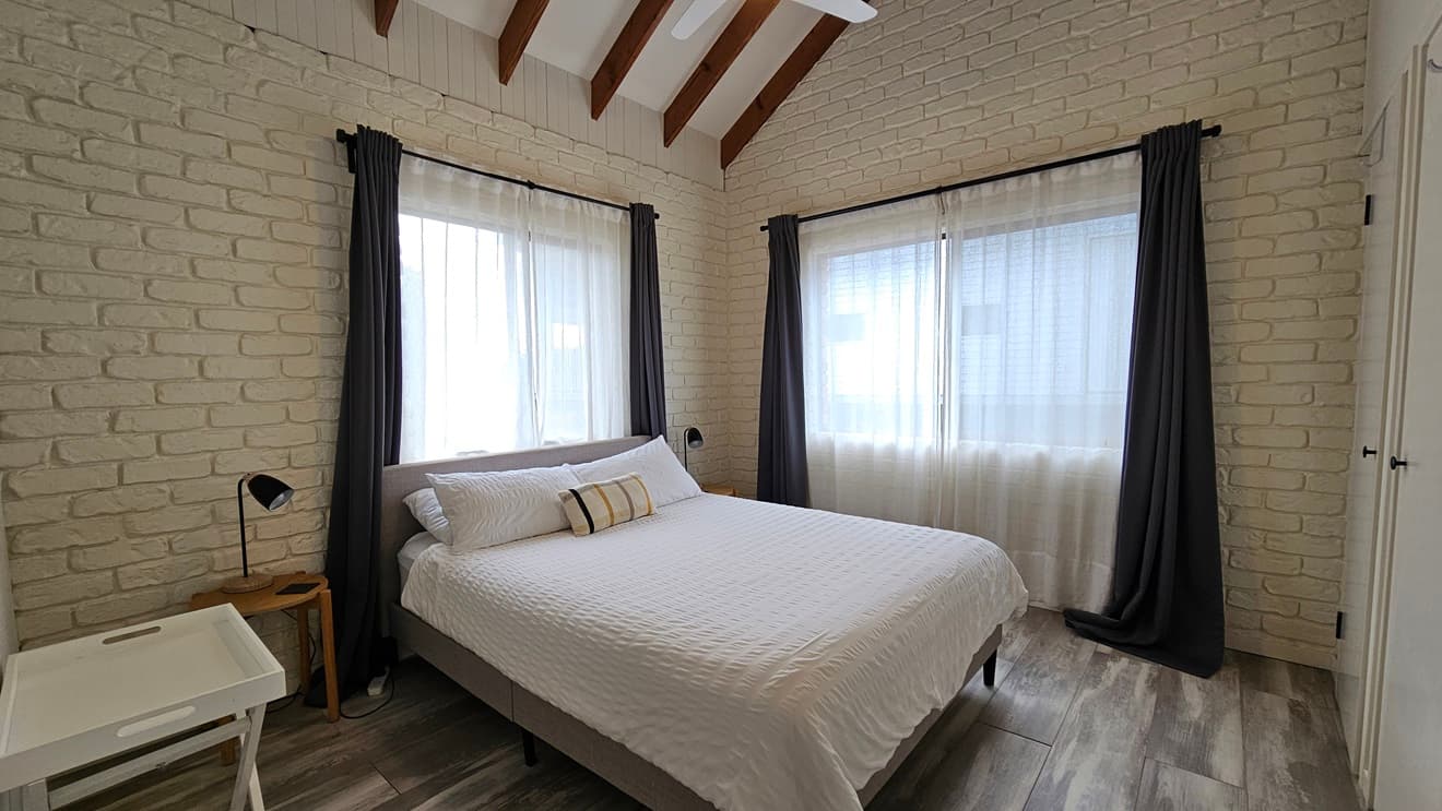 On the left, there is a bathroom with white brick walls, a small window, a round mirror, a sink, and a toilet. In the middle, there is a doorway leading to a bedroom with white brick walls, a bed with white bedding, and a large window with sheer curtains and dark drapes. On the right, there is a partial view of another doorway. The floor has laminate with a wooden design, and the ceiling is white with exposed wooden beams and a ceiling fan.