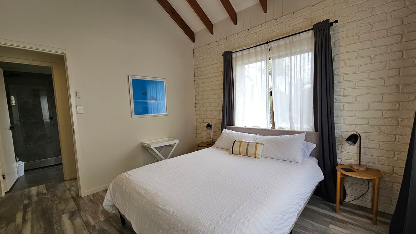 On the left, there is a doorway leading to a bathroom, with a white wall and a framed blue picture. In the middle, there is a bed with white bedding and a striped cushion, positioned against a white brick wall with a large window covered by sheer curtains and dark drapes. On the right, there is a small wooden side table with a black lamp. The floor has laminate with a wooden design, and the ceiling has exposed wooden beams.