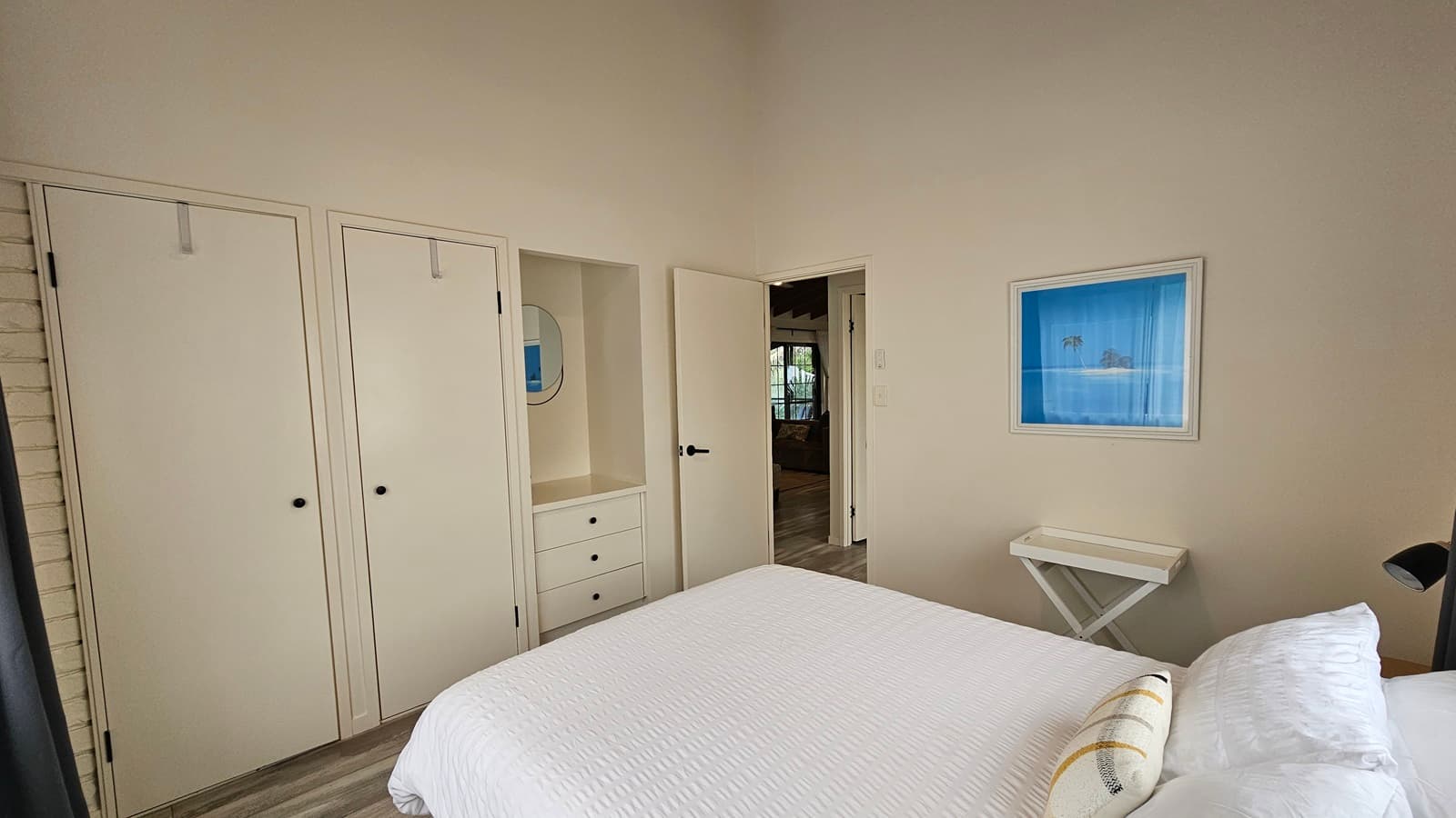 On the left, there are two white wardrobe doors with black knobs and a small built-in dresser with drawers. In the middle, there is a white door leading to another room, partially open, revealing a glimpse of a room with a large window and dark curtains. On the right, there is a white wall with a framed blue picture and a small white folding table. The floor has a light wood laminate design, and the ceiling is white and sloped.