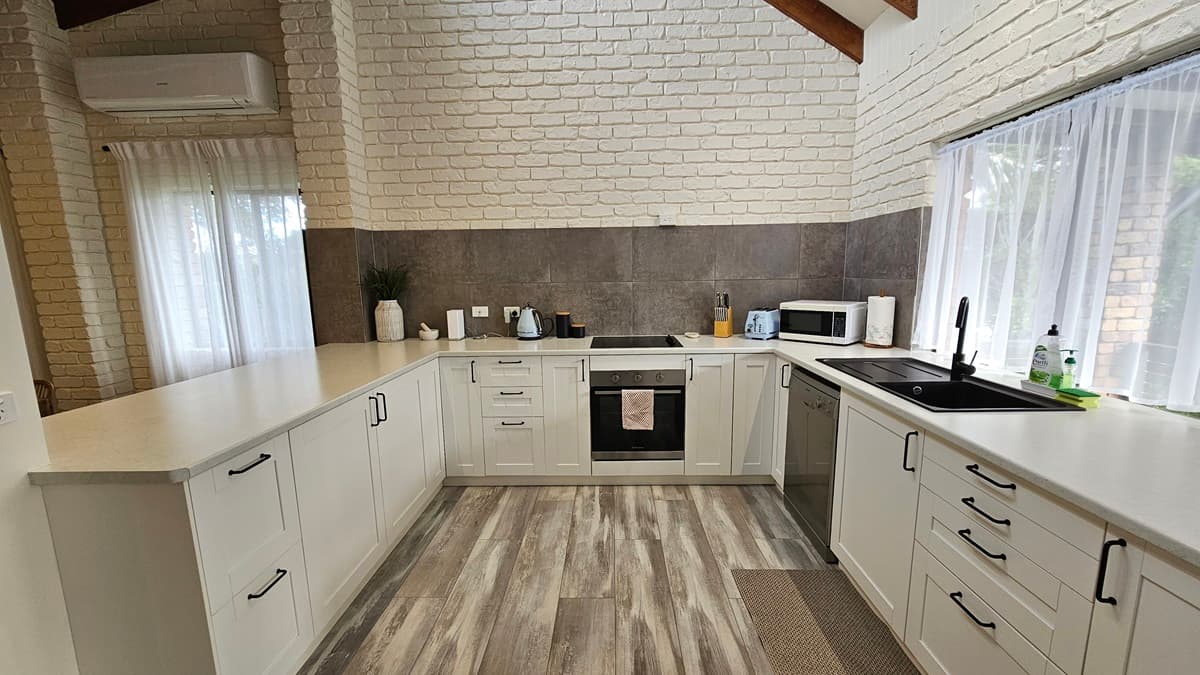 On the left, there is a stainless steel refrigerator and a living area with a brown sofa, a painting on the wall, and large windows with sheer curtains. In the middle, there is a white kitchen with cabinets, a sink, and a countertop. On the right, there is a white brick wall, a stove, a microwave, and a kettle. The floor has a wooden design laminate with a grey finish, and the ceiling has timber slats with ceiling fan and recessed lighting.