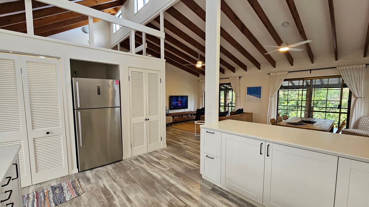 On the left, there is a stainless steel refrigerator next to white louvred doors. In the middle, there is a living area with a TV on a wooden stand, a brown sofa, and large windows with sheer curtains. In the background, there is a loft area with a railing. On the right, there is a dining table with chairs, more large windows with sheer curtains, and a painting on the wall. The floor has wooden design laminate with a grey finish, and the ceiling has exposed wooden beams with ceiling fans.