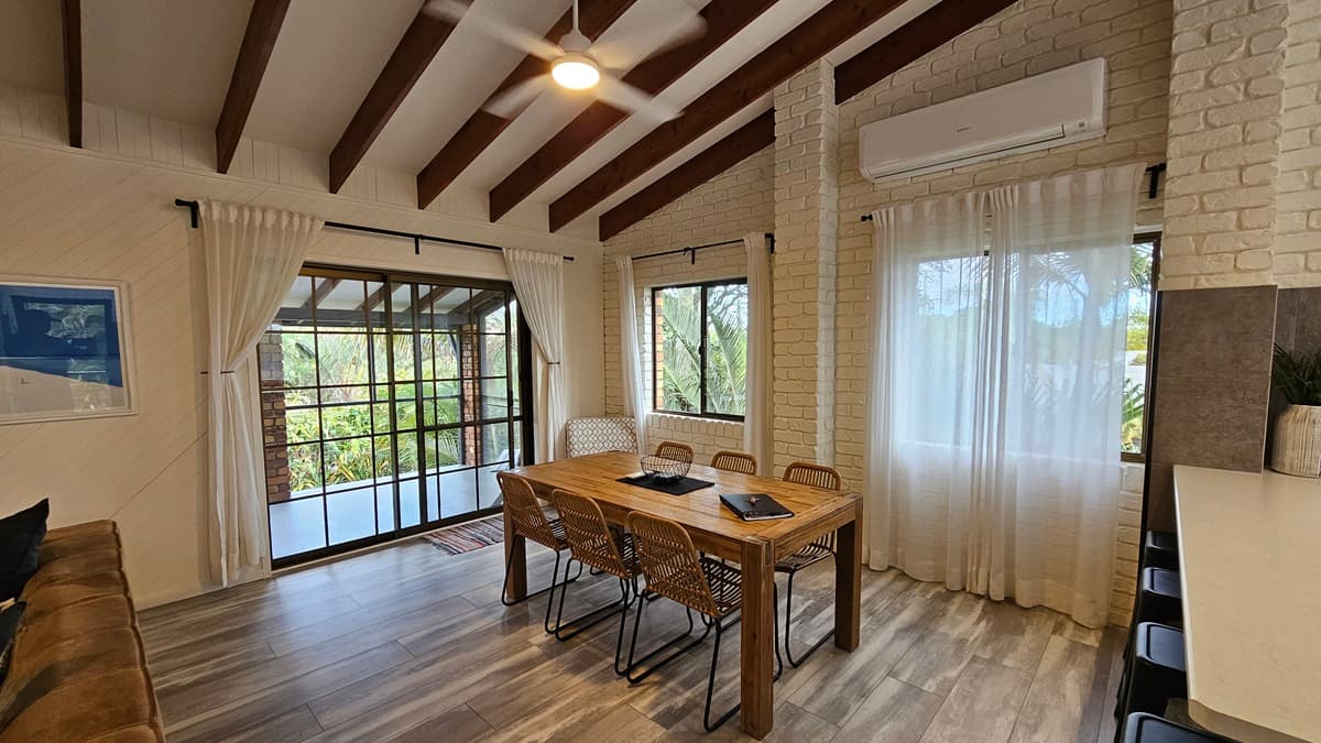 On the left, there is a black sofa with a framed picture above it and a large window with sheer curtains and a sliding glass door leading to a balcony. In the middle, there is a wooden dining table with four chairs and a laptop on the table. On the right, there is another large window with sheer curtains, a white brick wall, and an air conditioning unit above. The floor has wooden design laminate with a grey finish, and the ceiling has exposed wooden beams with a ceiling fan.