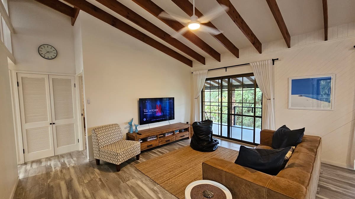 On the left, there are white louvred doors beneath a wall clock and a patterned armchair. In the middle, there is a wooden TV stand with a flat-screen TV and a large brown rug on the floor. There is a large window with sheer curtains and a sliding glass door leading to a balcony with a view of greenery. On the right, there is a brown sofa with cushions, a black bean bag, and a framed picture on the wall. The floor has wooden design laminate, and the ceiling has timber slats and a ceiling fan.