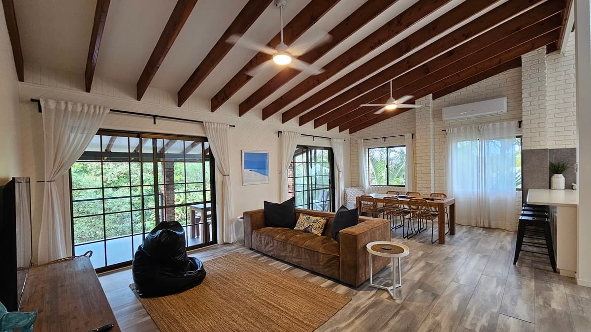 On the left, there is a window with sheer curtains and a glass door leading to a balcony with a view of trees. In the middle, there is a brown sofa with cushions, a small round side table, and a black bean bag on a brown rug. There is a dining table with chairs and another large window with sheer curtains. On the right, there is a white brick wall, a kitchen counter with stools, and an air conditioning unit above. The floor has laminate with a wooden design, and the ceiling has timber slats.