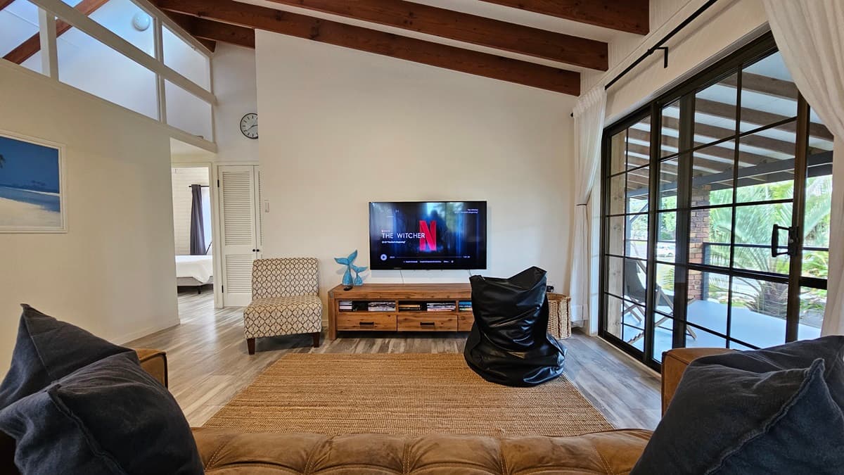 On the left, there is a patterned armchair next to white louvred doors and a wall clock above. In the middle, there is a wooden TV stand with a flat-screen TV and a black bean bag on a large brown rug. In the background, there is a glimpse of a bedroom through an open doorway. On the right, there is a large window with sheer curtains and a sliding glass door leading to a balcony with a view of palm trees. The floor has laminate with a wooden design and the ceiling has timber slats.