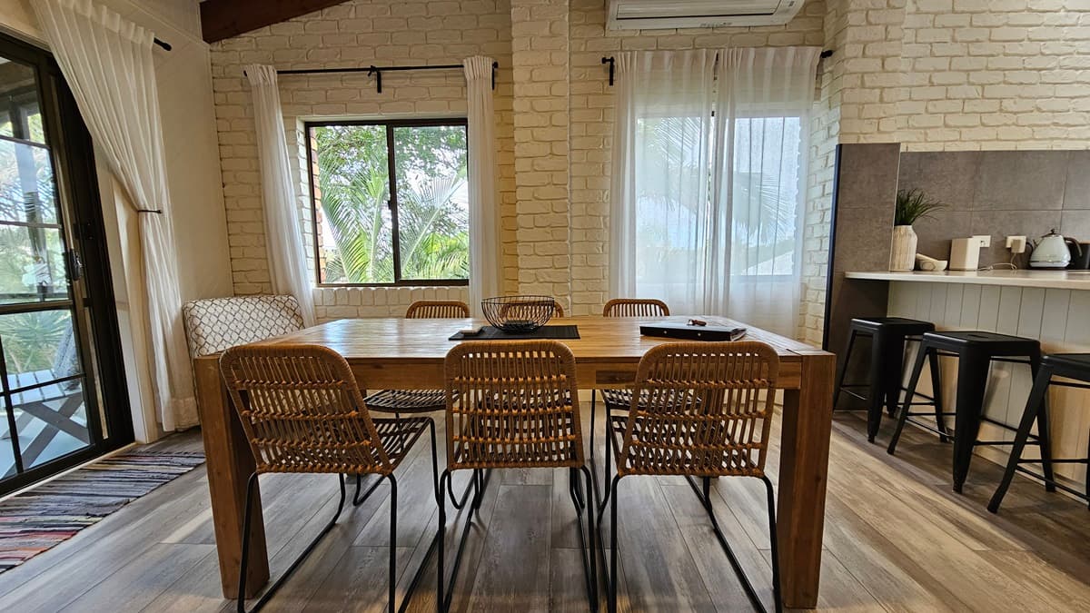 On the left, a sliding glass door with sheer curtains leads to a balcony. A patterned armchair sits next to a large window with sheer curtains. In the middle, there is a wooden dining table with six wicker chairs. On the right, another large window with sheer curtains, a white brick wall, and a kitchen counter with stools, a kettle, and a toaster. The floor has a wooden design, and the ceiling features exposed wooden beams and timber slats.