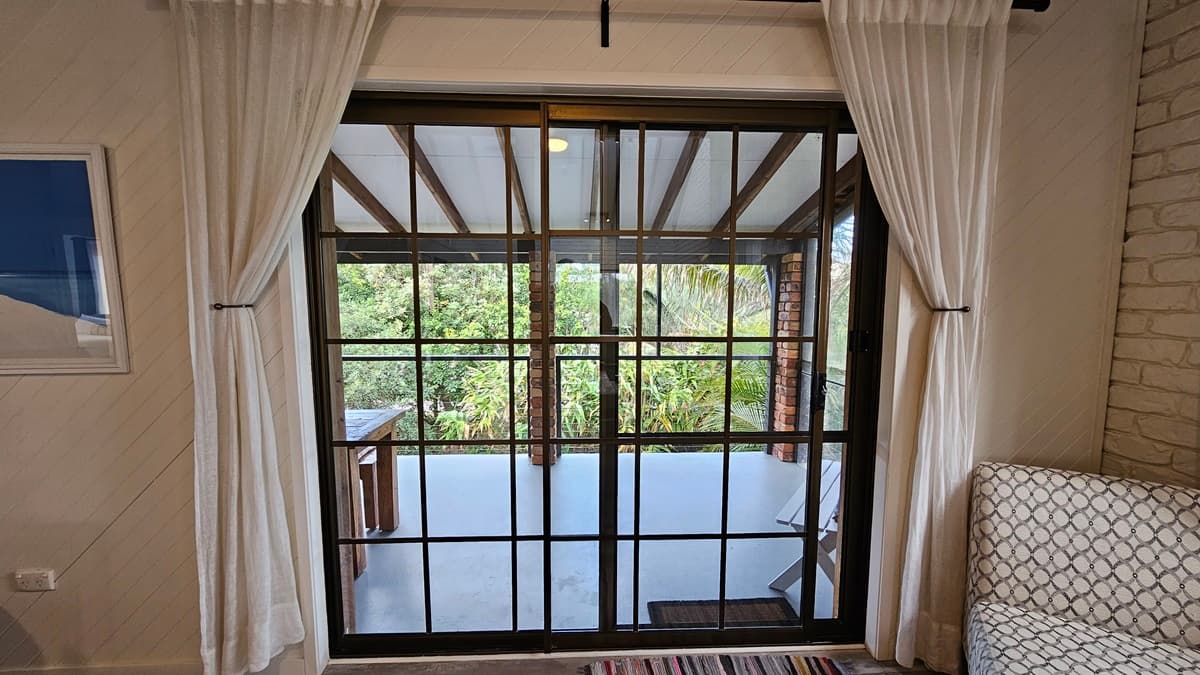 On the left, there is a framed picture on the wall and a white curtain partially covering a sliding glass door. In the middle, there is a large sliding glass door with black frames leading to a balcony with a view of trees and greenery. On the right, there is a white brick wall and a patterned armchair. The floor has laminate with a wooden design, with a striped rug in front of the door, and the ceiling has timber slats
