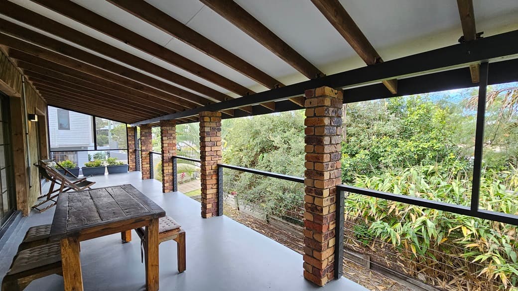 On the left, there is a wooden table with benches and two wooden chairs against a wall. In the middle, there are brick columns supporting the roof, with black metal railings and glass panels between them. On the right, there is a view of dense greenery, including bamboo and other trees. The floor is a smooth, light grey surface, and the ceiling has exposed wooden beams with white panels in between.