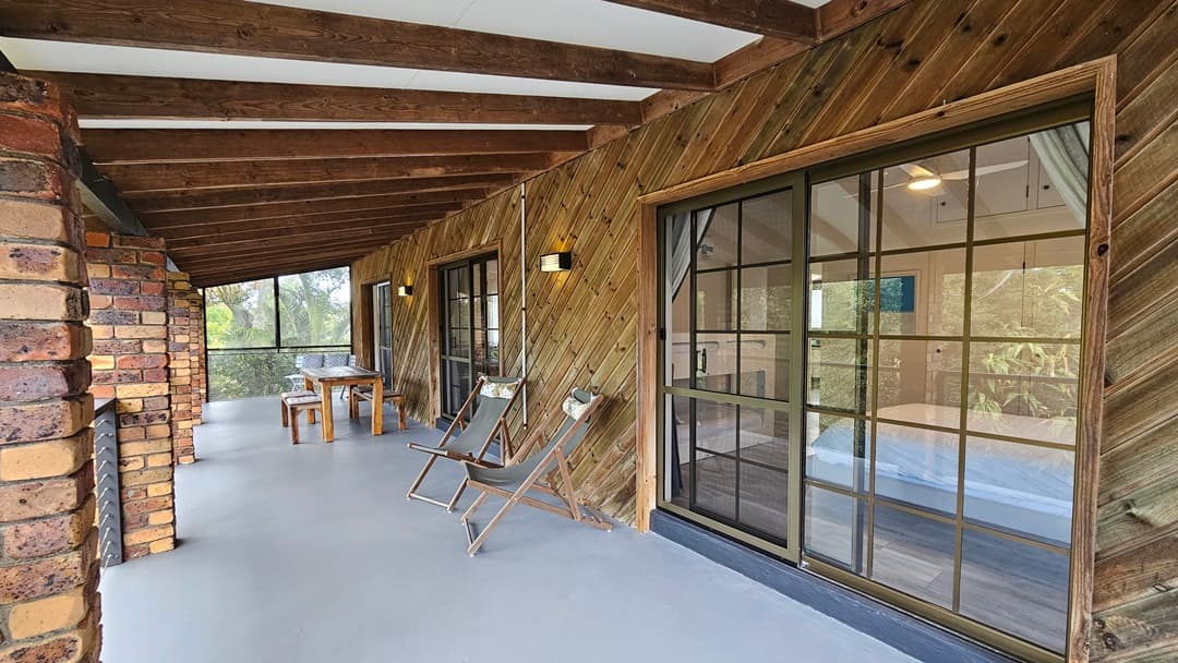 On the left, there is a brick column and a wooden table with benches. In the middle, there are two wooden deck chairs facing a sliding glass door with a black frame. On the right, the sliding glass door reveals an interior room with a bed and a ceiling fan. The background features a wooden wall with diagonal planks and wall-mounted lights. The floor is a smooth grey surface, and the ceiling has exposed wooden beams with white panels in between.