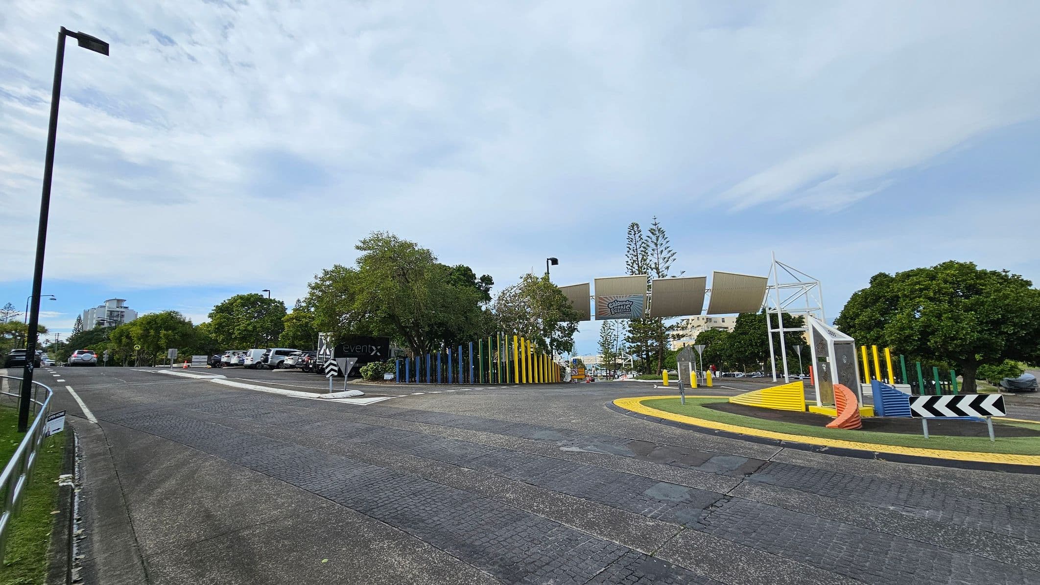 A road with a gentle curve leads to a parking area on the left, where several cars are parked. A tall streetlamp stands on the left side of the road. On the right, there is a colourful installation with vertical poles in various colours and a structure with angled panels. Trees are scattered throughout the area, and a building is visible in the distance. The road surface is textured, and there are directional signs near the installation.