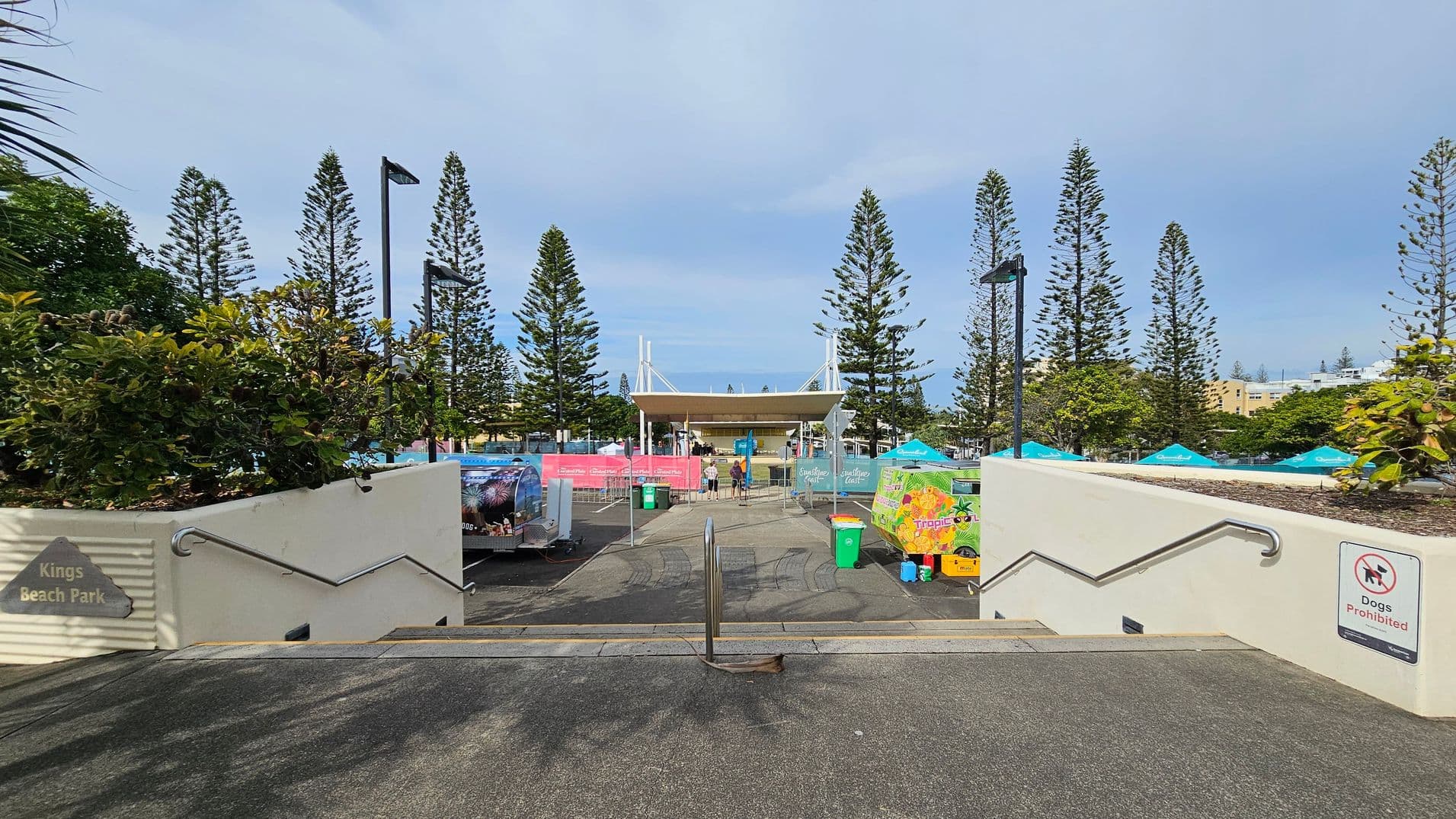 A pathway leads downwards with handrails on both sides. Signs indicate "Kings Beach Park" and "Dogs Prohibited". In the background, a pavilion is visible, surrounded by tall pine trees. There are colourful banners and umbrellas, along with bins and a few people near the pavilion. The ground is paved, and the area is well-lit with street lamps.