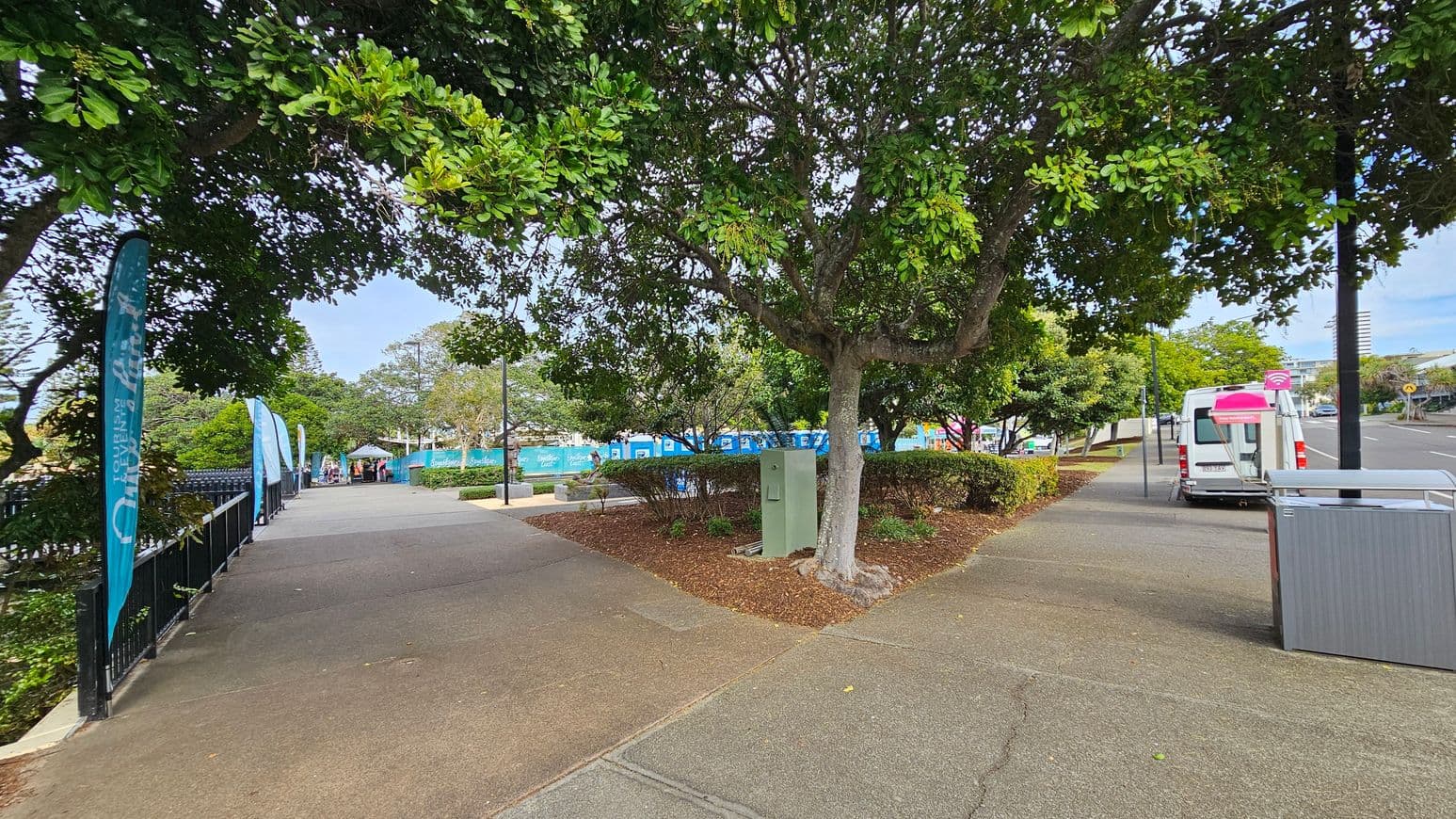 A paved walkway lined with blue banners on the left, bordered by a black metal fence. Trees with dense green foliage provide shade. On the right, a white van with a pink roof is parked on the street. A green utility box and a rubbish bin are near the tree. In the background, more trees and a few people are visible near a blue construction barrier. The ground is a mix of concrete and mulch, with some grass patches.