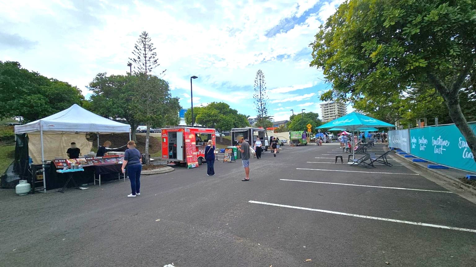 A market scene with a white tent on the left, where two people are serving food. Next to it is a red food truck with a person standing in front. Further along, there are more food stalls and a few people walking. On the right, there are chairs and tables under a large turquoise umbrella. Trees line the background, and a tall building is visible in the distance. The ground is paved, and there are parking lines visible.