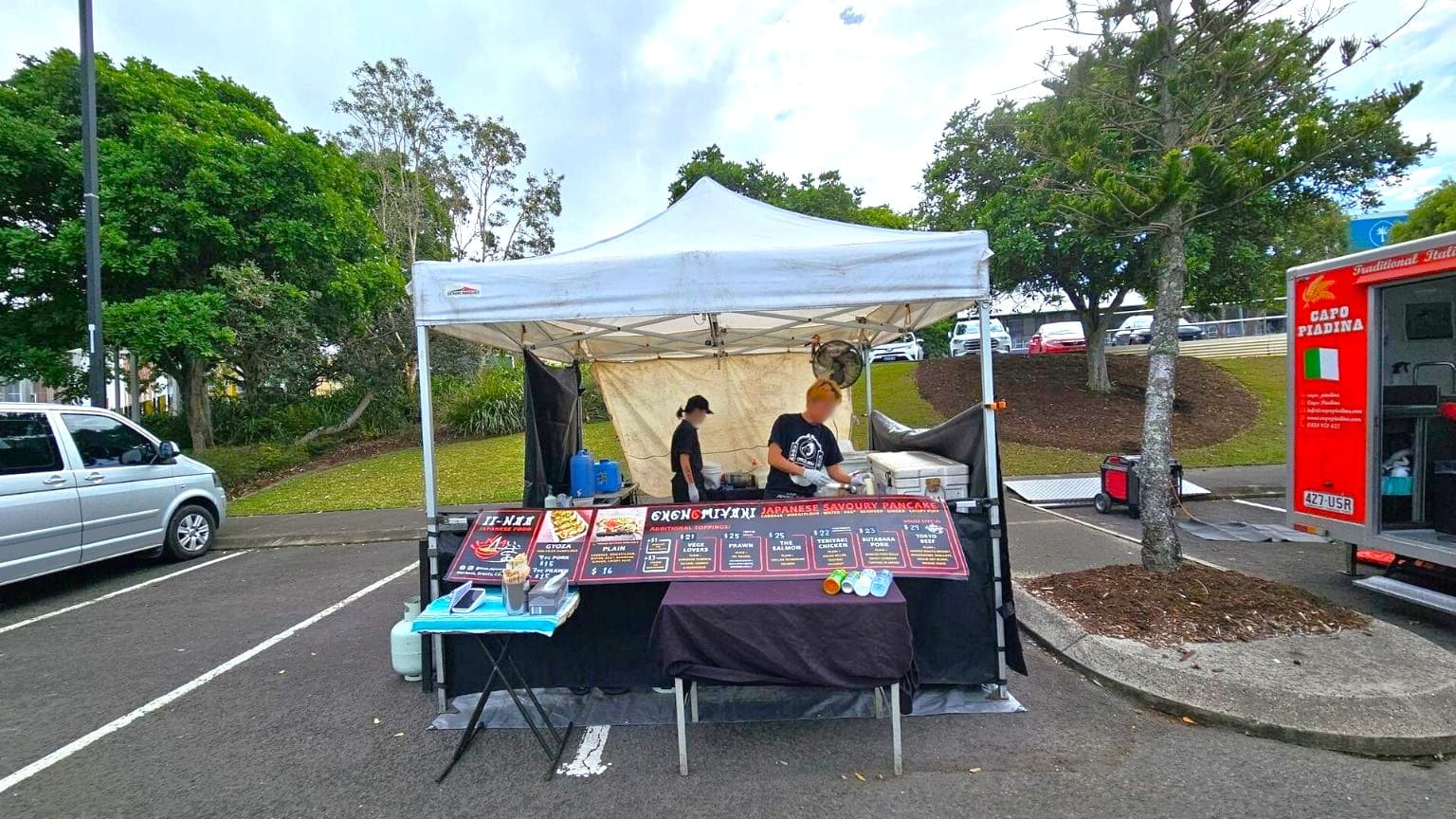 A food stall with a white canopy is set up in a car park. In the middle, two people are preparing food behind a counter displaying a menu for Japanese savoury pancakes. On the left, a silver van is parked. On the right, a red food truck with Italian branding is visible. The background features a grassy area with trees and parked cars. The ground is asphalt with marked parking spaces.