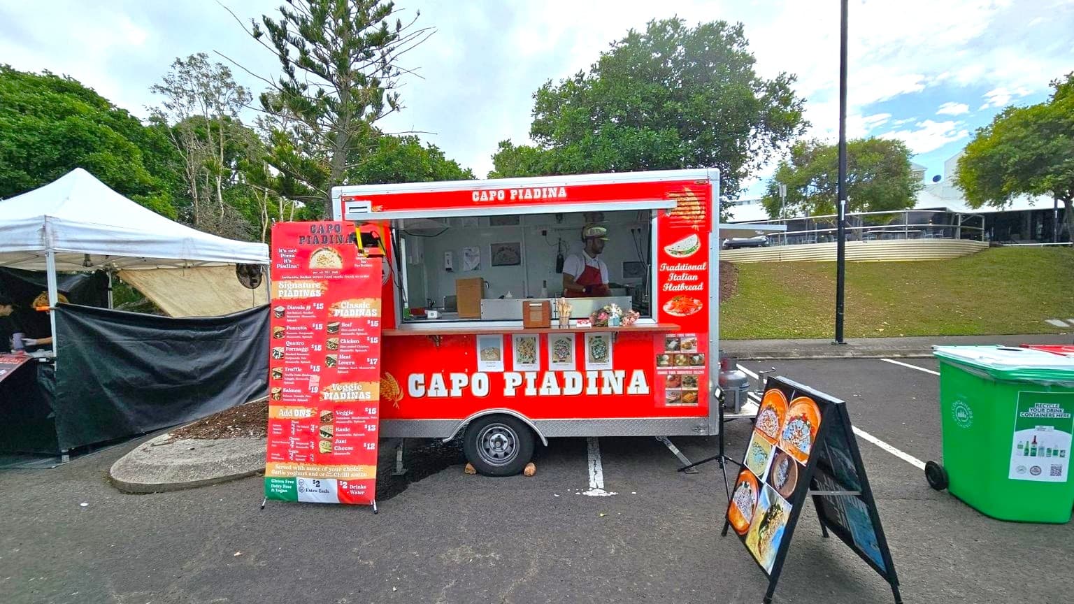 A red food truck labelled "Capo Piadina" is in the centre, offering traditional Italian flatbread. A menu board stands to the left, listing various piadina options. A person is visible inside the truck. To the right, a green recycling bin is present. In the background, there are trees and a grassy area with a building and a railing. A white tent is partially visible on the left. The ground is paved, and a blackboard with images of food is in front of the truck.