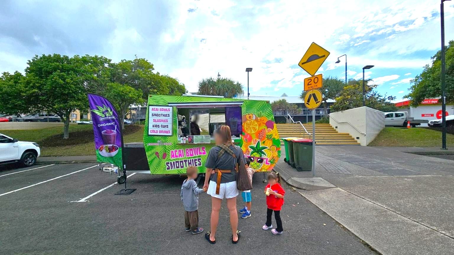 A food truck with vibrant fruit-themed graphics is in the centre, offering acai bowls and smoothies. A woman with three children stands in front, with one child holding a drink. To the right, a yellow speed limit sign and a pedestrian crossing sign are visible. Behind the truck, there are stairs leading up to a building, and trees are on the left. A white vehicle is parked on the left side of the image. The ground is paved, and there are bins near the truck.