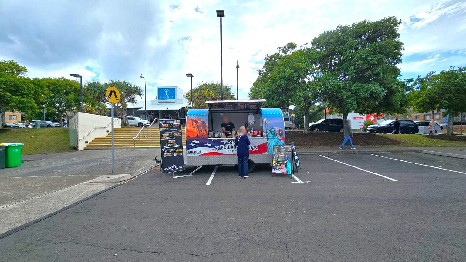 A food truck is parked in an empty car park, with a person standing in front of it. The truck has colourful graphics featuring American landmarks. Two menu boards are displayed outside. In the background, there are trees, a building with a sign, and a few parked cars. A pedestrian is walking on the right side. The ground is asphalt, and there are marked parking spaces. A set of stairs and a green bin are visible on the left.