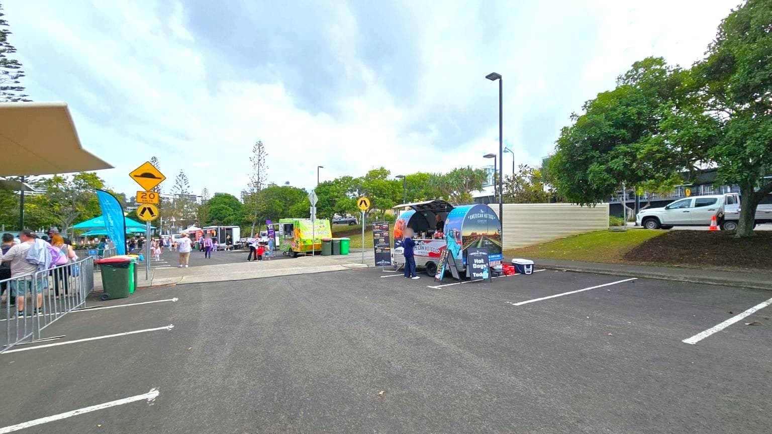 A street market scene with food stalls and people. On the left, a group of people stand near a barrier and a green bin, with a blue flag and a canopy visible. In the centre, a yellow road sign indicates a speed bump and a 20 km/h limit. In the middle, a food truck with "American Hotdogs" signage is attended by a person. On the right, a white vehicle is parked near trees and a corrugated fence. The ground is asphalt, and the sky is overcast.