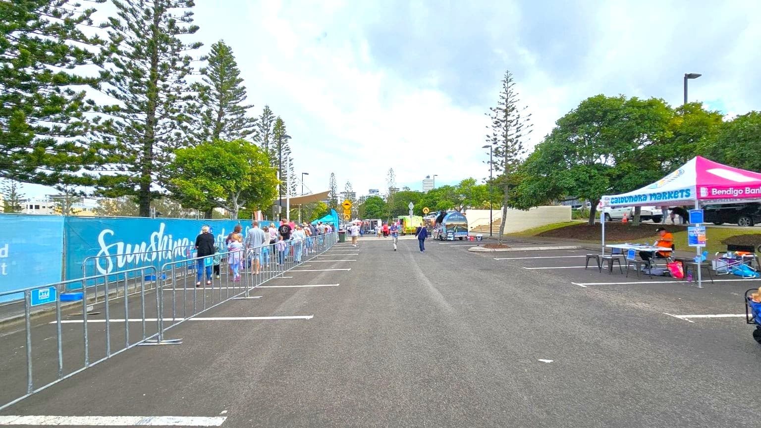 A long queue of people is lined up on the left side, separated by metal barriers. Tall trees and a blue fence with text are behind them. On the right, a pink and white tent labelled "Caloundra Markets" and "Bendigo Bank" has a person sitting at a table with various items. The background features more trees and a few distant buildings. The ground is an asphalt surface with parking lines visible.