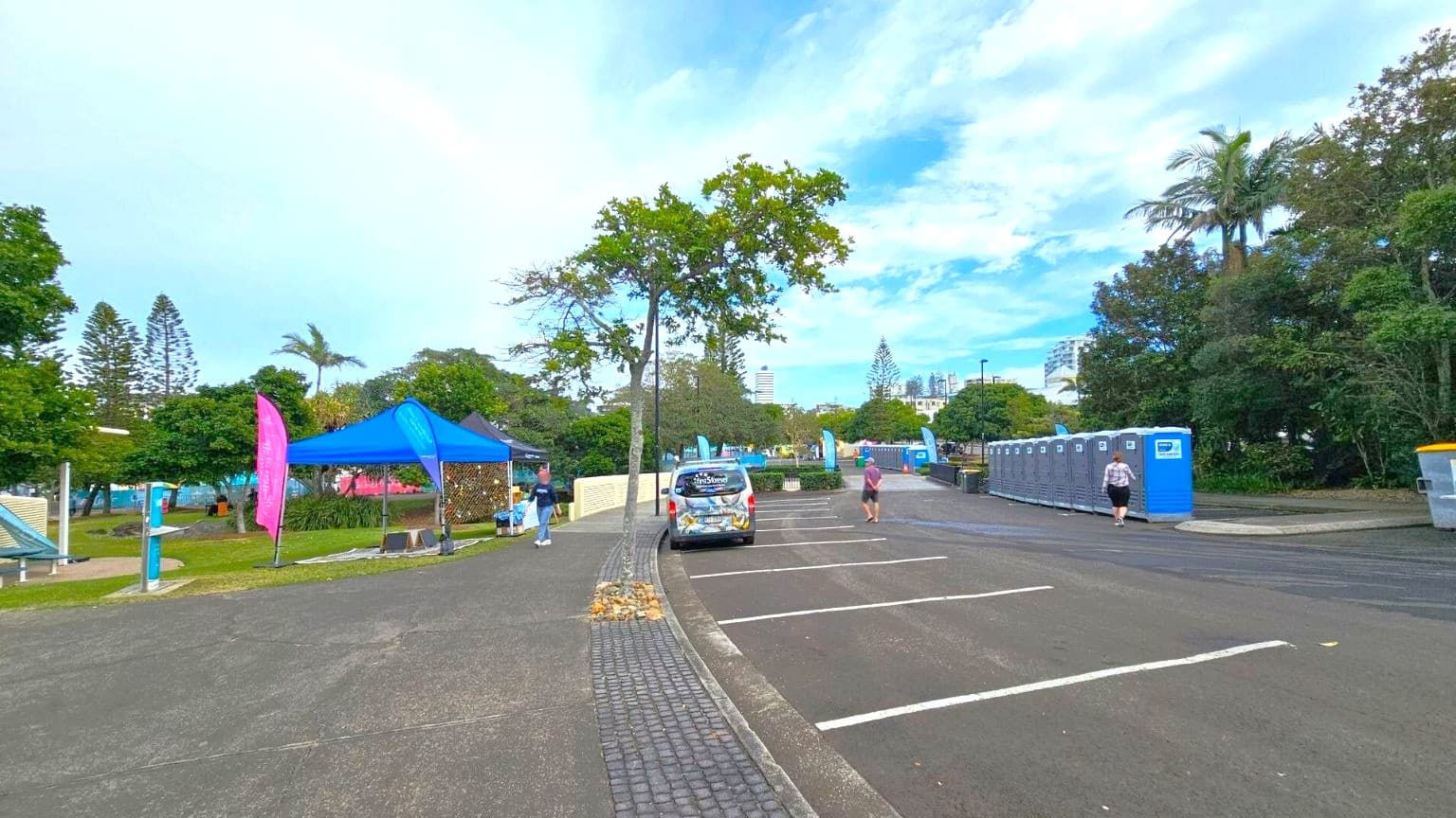A park setting with a paved path in the centre. On the left, there's a blue tent with a pink flag nearby, surrounded by trees and grass. A car is parked on the path. On the right, a row of portable toilets is lined up next to more trees. A person is walking towards the toilets. In the background, there are more trees and a glimpse of buildings. The sky is partly cloudy.