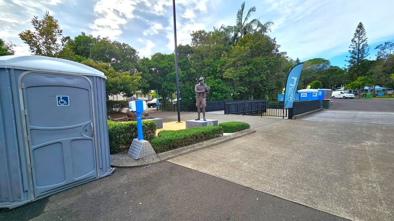 A portable toilet with a wheelchair symbol is on the left. In the centre, a statue of a soldier stands on a pedestal surrounded by low hedges. A tall flagpole is nearby. On the right, a blue flag with "Queensland" written on it is next to a blue container. Trees and a parked white van are in the background. The ground is a mix of asphalt and concrete.