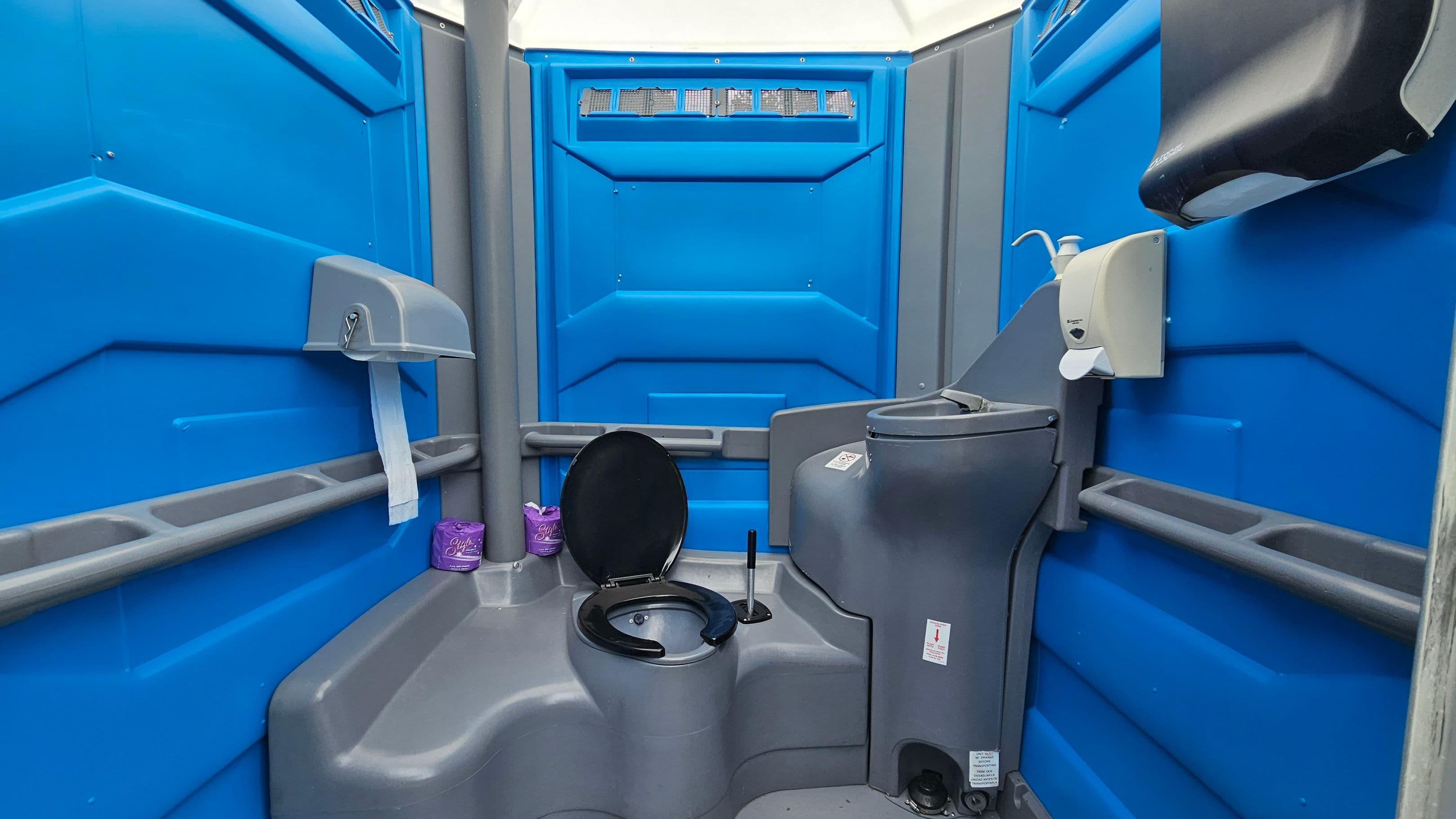 Inside a portable toilet with blue walls. On the left, a toilet paper dispenser with a roll of paper hanging down. Below, two purple toilet paper rolls are placed on a ledge. In the centre, a black toilet seat is open. On the right, a grey sink with a soap dispenser above it and a paper towel dispenser mounted on the wall. The ceiling is translucent, allowing light to enter. The floor is grey with a textured surface.