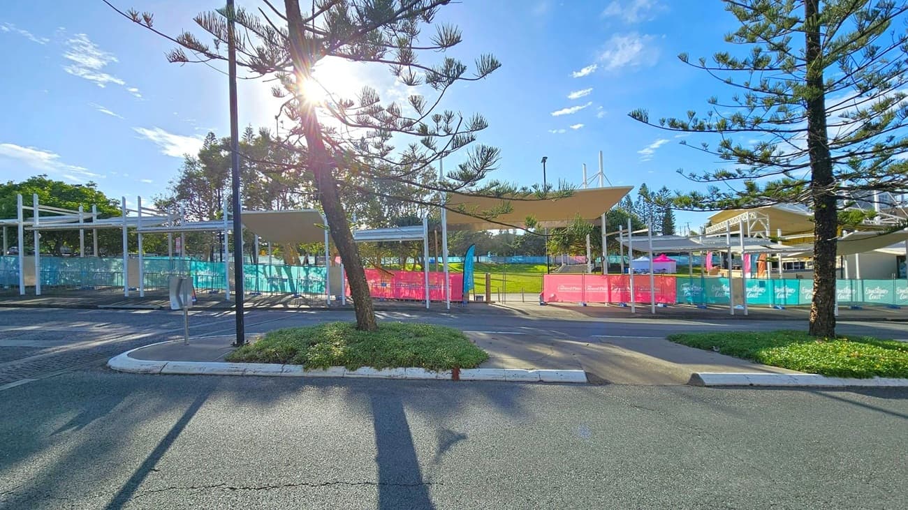 A paved area with a few tall, slender trees casting long shadows on the ground. In the background, there are white metal structures with shade sails, surrounded by colourful fencing in pink and blue. The sun is shining brightly through the trees, creating a lens flare effect. The ground is asphalt with a small patch of greenery near the centre.