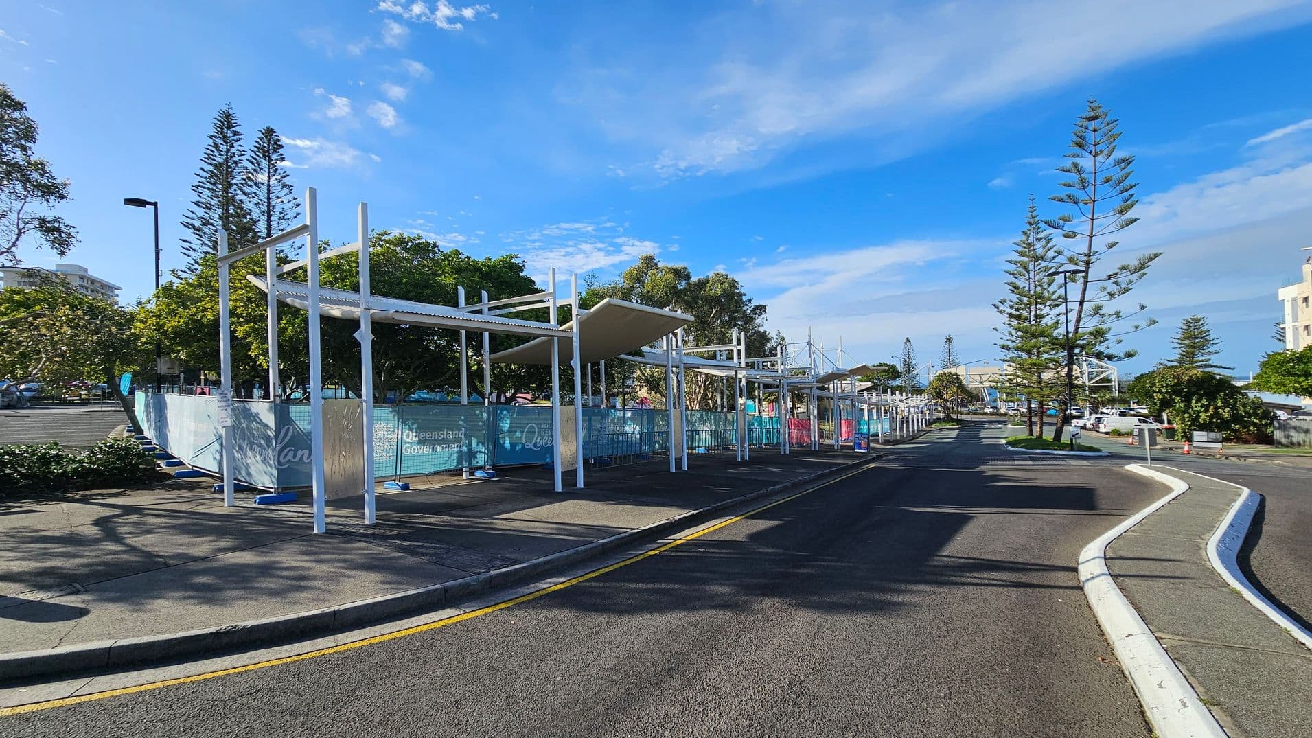 A street scene with a covered walkway on the left, featuring white metal structures and a corrugated roof. Blue fencing with "Queensland Government" branding lines the area. Tall, slender trees are visible in the background, along with a few parked cars. The road curves to the right, bordered by a white kerb and a narrow strip of grass. Buildings and more trees are visible in the distance under a clear blue sky.