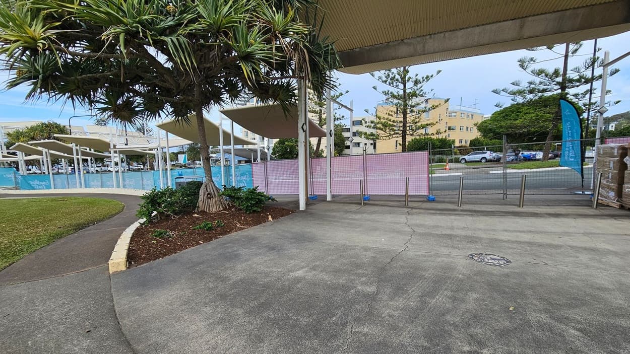A paved area with a tree and some plants on the left. In the middle, there are several white structures with angled roofs, and a pink mesh fence runs along the perimeter. In the background, a yellow building and parked cars are visible. The ground is concrete, and the ceiling is not visible.