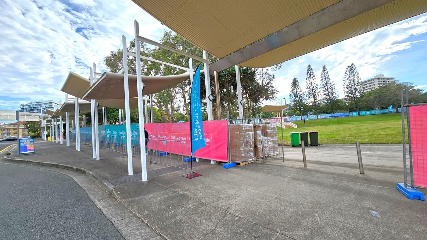 A paved walkway with a series of white metal structures and beige canopies on the left. Blue and pink banners are attached to fencing, with stacks of boxes behind them. A grassy area with trees and a few buildings is visible on the right. A couple of green bins are near the grass. The ground is concrete, and the sky is partly cloudy.