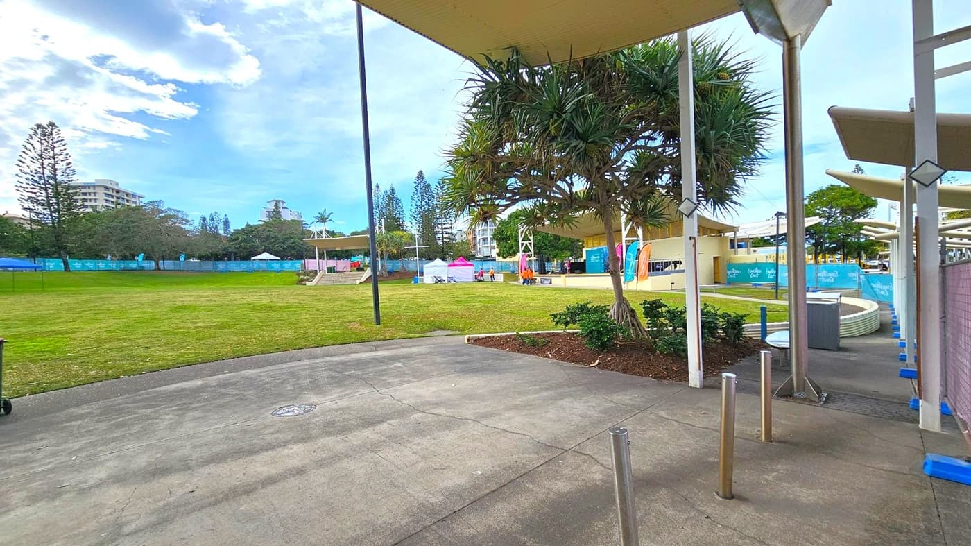 A grassy area with a concrete path in the foreground. On the right, there are metal poles and a covered walkway with a tree nearby. In the middle, several tents and banners are set up, with people gathered around. The background features tall trees and buildings, partially obscured by a fence with blue banners. The sky is partly cloudy.