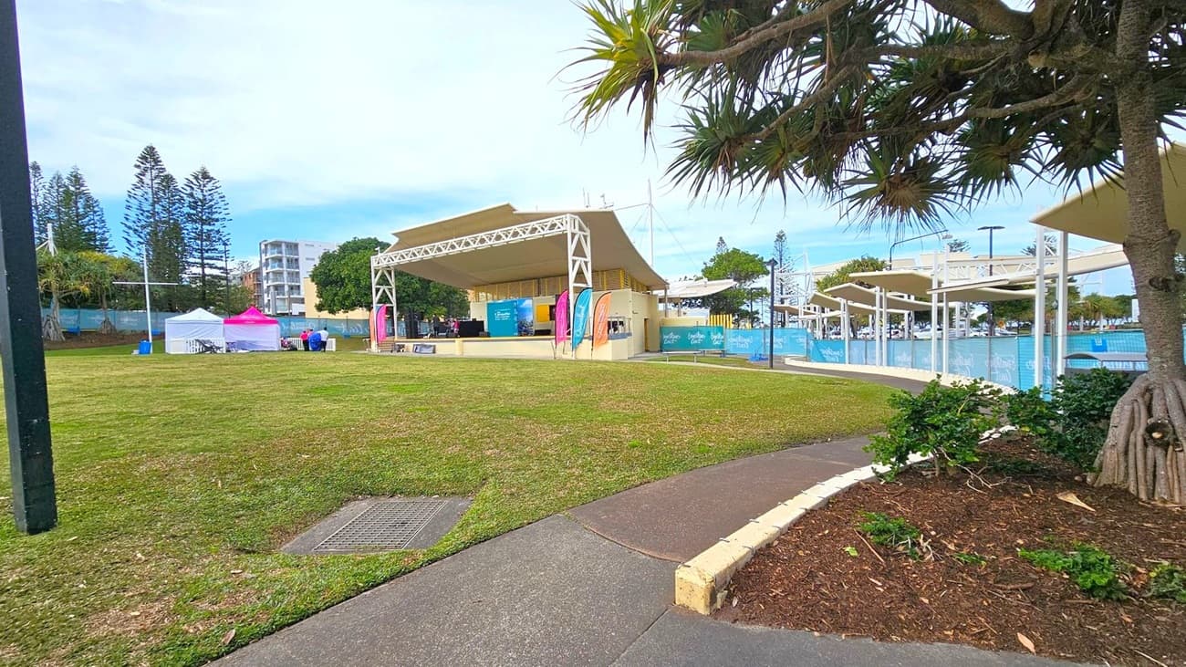 A grassy area with a paved path in the foreground. On the right, a tree with exposed roots and some small plants. In the centre, a stage with a canopy and colourful banners. To the left, two tents, one white and one pink, with people sitting nearby. In the background, tall trees and a multi-storey building. The sky is overcast.