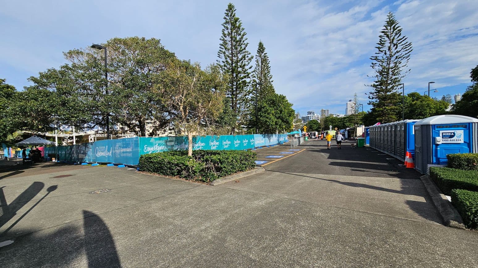 A paved pathway with portable toilets lined up on the right side, marked by a blue and white sign. Two people are walking along the path. On the left, there are tall trees and a blue fence with "Queensland Government" and "Sunshine Coast" branding. The background features more trees and distant buildings. The ground is a mix of concrete and green patches, and there are shadows cast by nearby objects.