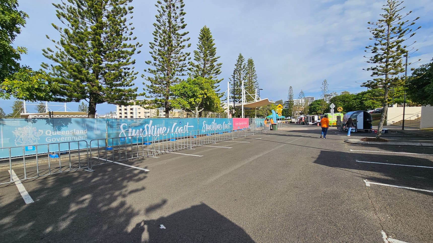 Tall pine trees line both sides of a paved area, with metal barricades forming a pathway. On the left, a blue banner with "Queensland Government" and "Sunshine Coast" text is visible. In the background, there are buildings and a white structure with a canopy. On the right, two people walk towards a small tent, with more trees and a few parked vehicles nearby. The ground is marked with parking lines, and shadows from the trees are cast on the pavement.