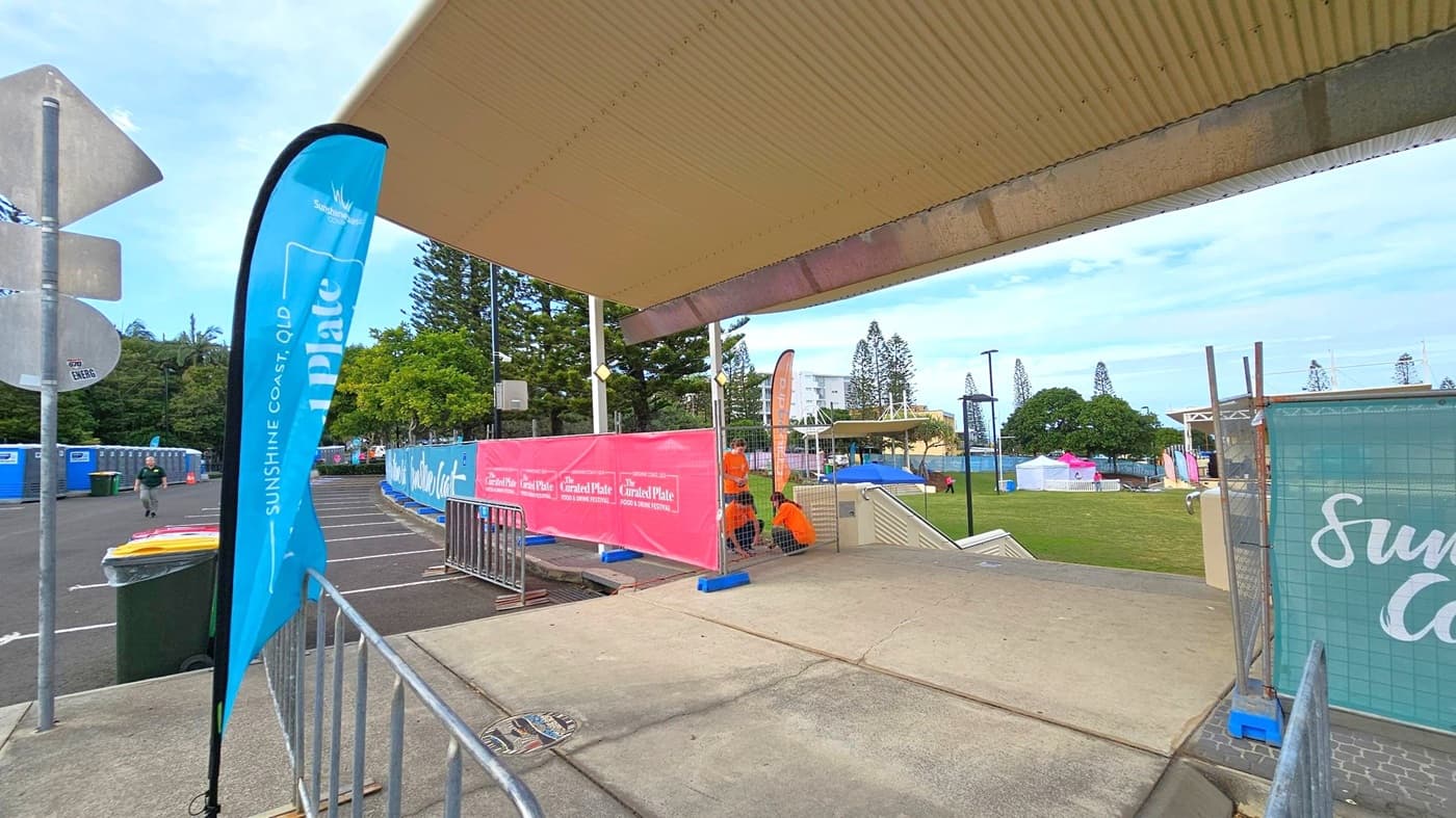 A covered walkway with a corrugated roof leads to an open area. On the left, a blue flag reads "Sunshine Coast, QLD" next to a bin and a few signs. In the middle, a pink banner advertises "The Curated Plate" food festival. Two people in orange shirts are near the banner. On the right, a grassy area with tents and trees is visible. The ground is a mix of concrete and grass, and there are metal barriers along the path.