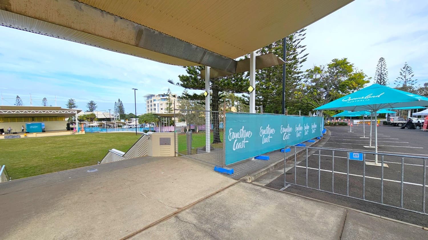 A grassy area is on the left with a stage and a few people in the distance. On the right, there are blue umbrellas with "Queensland Australia" written on them, set up over a paved area. A barrier with "Sunshine Coast" branding separates the two sections. In the background, there are trees and a multi-storey building. The ceiling is a corrugated structure, and the floor is a mix of concrete and grass.