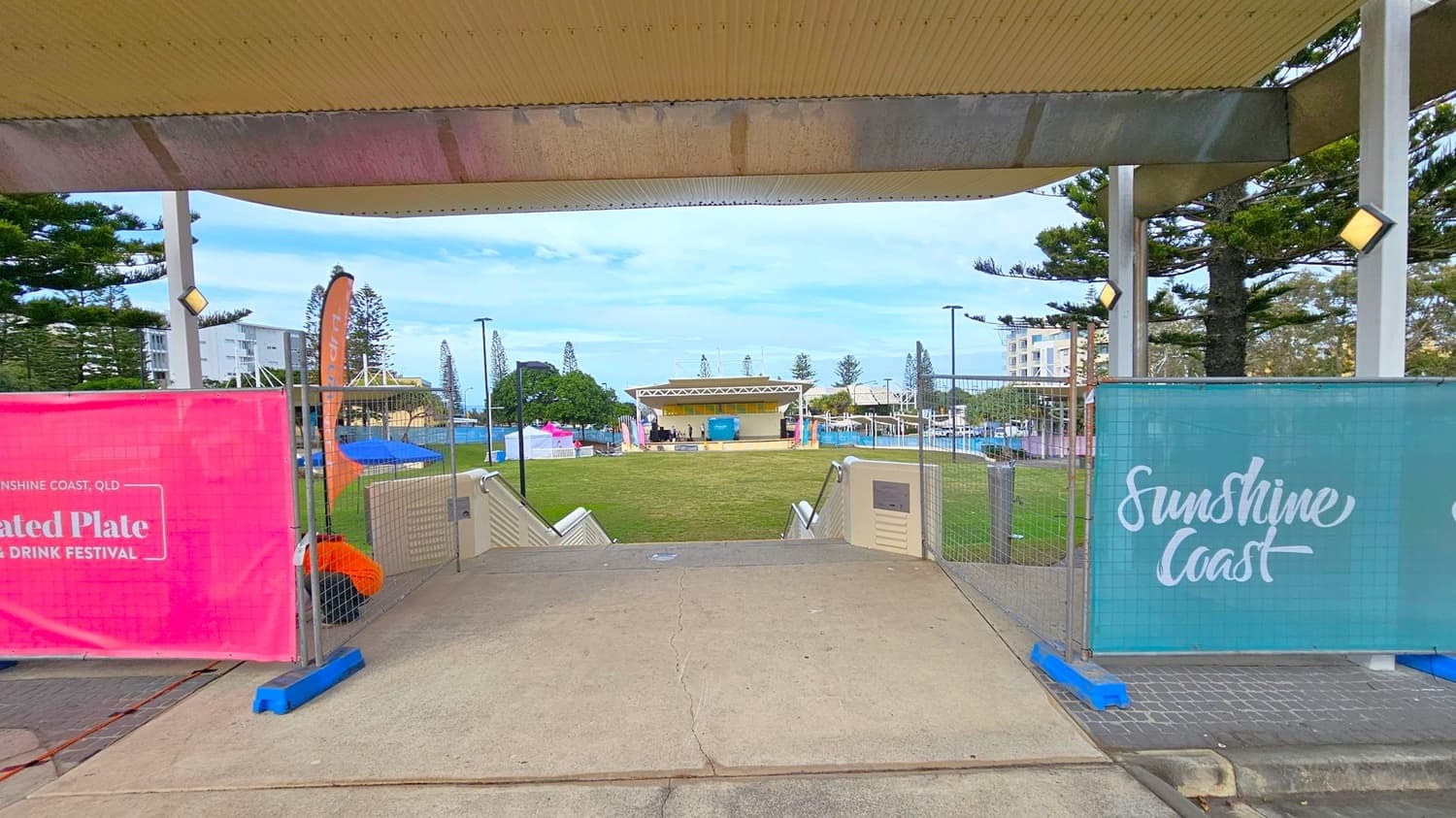 Under a corrugated ceiling, a pathway leads to a grassy area with a stage in the centre. On the left, a pink banner advertises a food and drink festival, while on the right, a blue banner reads "Sunshine Coast." Tall trees and a few buildings are visible in the background. The area is fenced, with steps leading down to the grass.