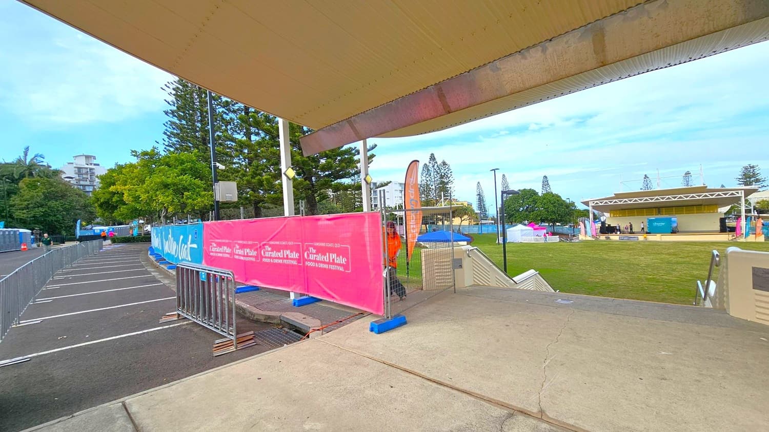 A covered walkway with a beige ceiling and concrete floor leads to an open grassy area. On the left, there are metal barriers and pink banners advertising "The Curated Plate Food & Drink Festival." Trees and a building are visible in the background. On the right, a stage with a white roof is set up on the grass, with several tents and flags nearby. A person in an orange shirt stands near the banners.