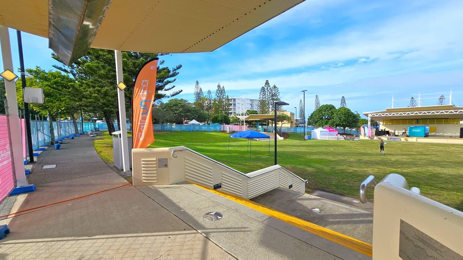 A park area with a concrete path on the left, lined with trees and a pink mesh fence. An orange flag with text is near the path. In the centre, a grassy field with a blue canopy and a person walking. On the right, a stage with a roof and some equipment. Buildings and more trees are visible in the background. The foreground has a ramp with a yellow tactile strip and metal railings.