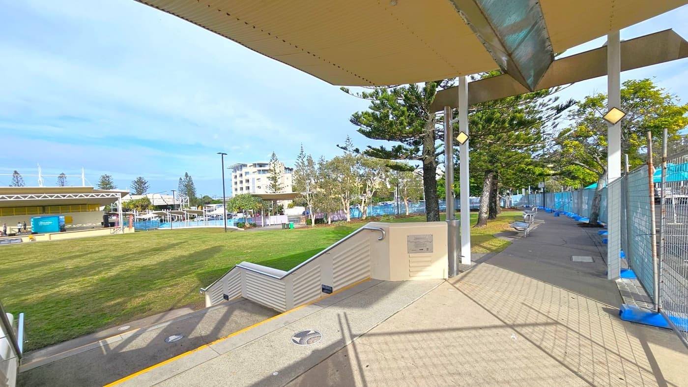 A covered walkway with a corrugated ceiling and metal supports on the right. The path is lined with a fence and several benches. On the left, a grassy area leads to a building with a flat roof and a blue sign. In the background, there are trees and a multi-storey building. The ground is a mix of concrete and grass.
