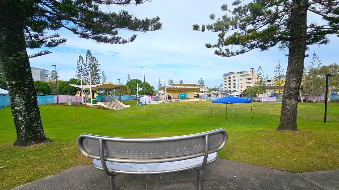 A park scene with a metal bench in the foreground, positioned between two large trees. The ground is covered with well-maintained grass. In the middle distance, there is a stage with a canopy and some colourful tents. Buildings are visible in the background, along with more trees and a few streetlights. A blue canopy tent is set up on the right side of the grassy area.