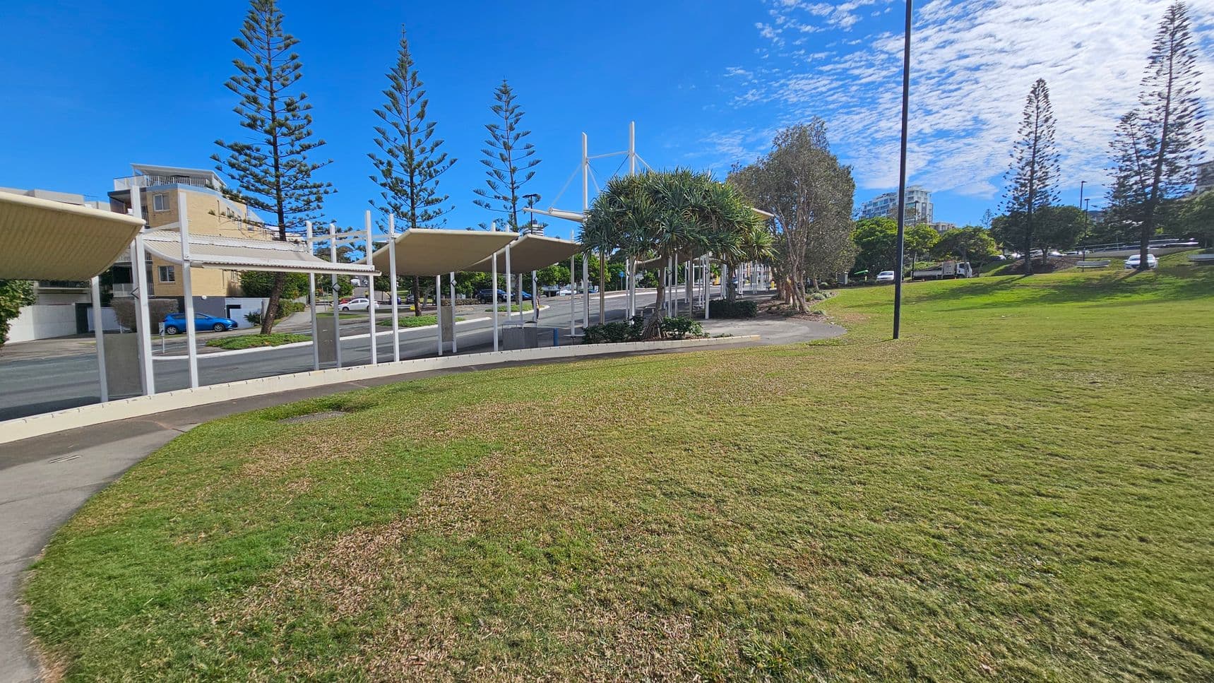 A grassy park area with patches of brown grass in the foreground. On the left, there are several covered walkways with white supports. Behind them, a street with a few parked cars and a beige building. In the middle, tall, slender trees are visible. On the right, more trees and a lamppost are present, with additional buildings partially visible in the background. The sky is clear with some clouds.