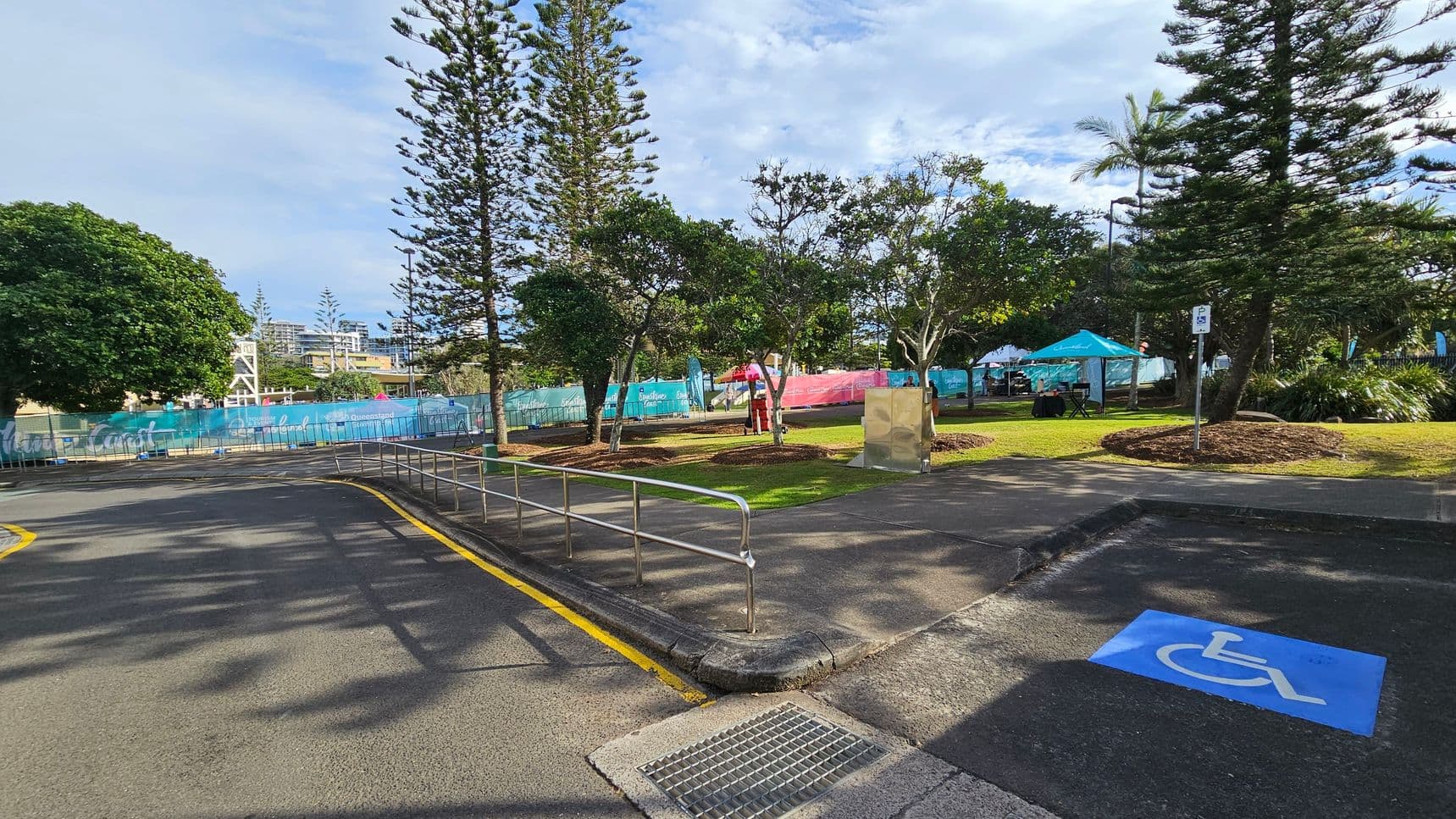 A paved area with a marked disabled parking space in the foreground. A metal railing lines the edge of the pavement. In the background, there are several tall trees and a grassy area with a few people standing near colourful banners and tents. A large tree is on the left, and a building with a clock tower is visible in the distance. The scene is well-lit, suggesting daytime.