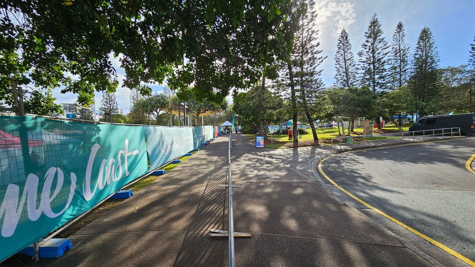 A pathway lined with a turquoise barrier on the left, featuring white text. The path is bordered by metal railings and leads towards a distant tent. On the right, a grassy area with tall trees and a black vehicle parked near a curved road. Shadows of the trees are cast on the pavement, and the area is well-lit by sunlight filtering through the foliage above.
