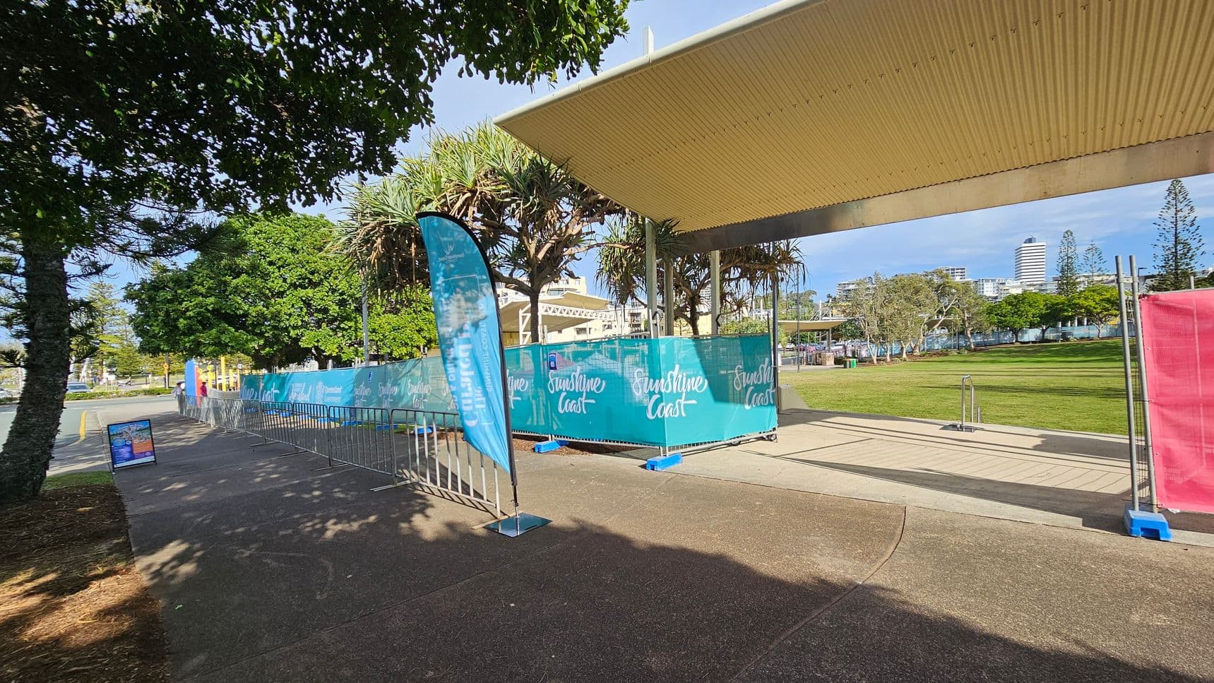 A pathway lined with metal barriers and blue banners reading "Sunshine Coast" on the left. Tall trees provide shade, and a signboard is visible near the path. On the right, a grassy area extends with scattered trees and a few buildings in the distance. A large, flat-roofed structure provides partial cover. The ground is paved, and the sky is clear.