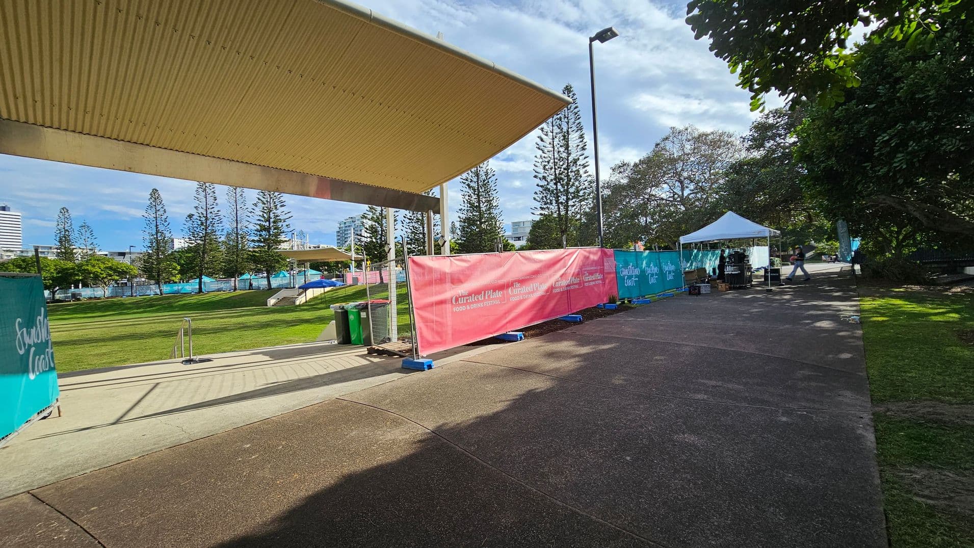 A pathway runs through a park area with a large canopy structure on the left. Pink and teal banners with "Curated Plate Food & Drink Festival" line the path. A white tent is set up on the right with equipment underneath. Tall trees and a few buildings are visible in the background. The ground is a mix of grass and concrete, and there are some shadows from the trees.